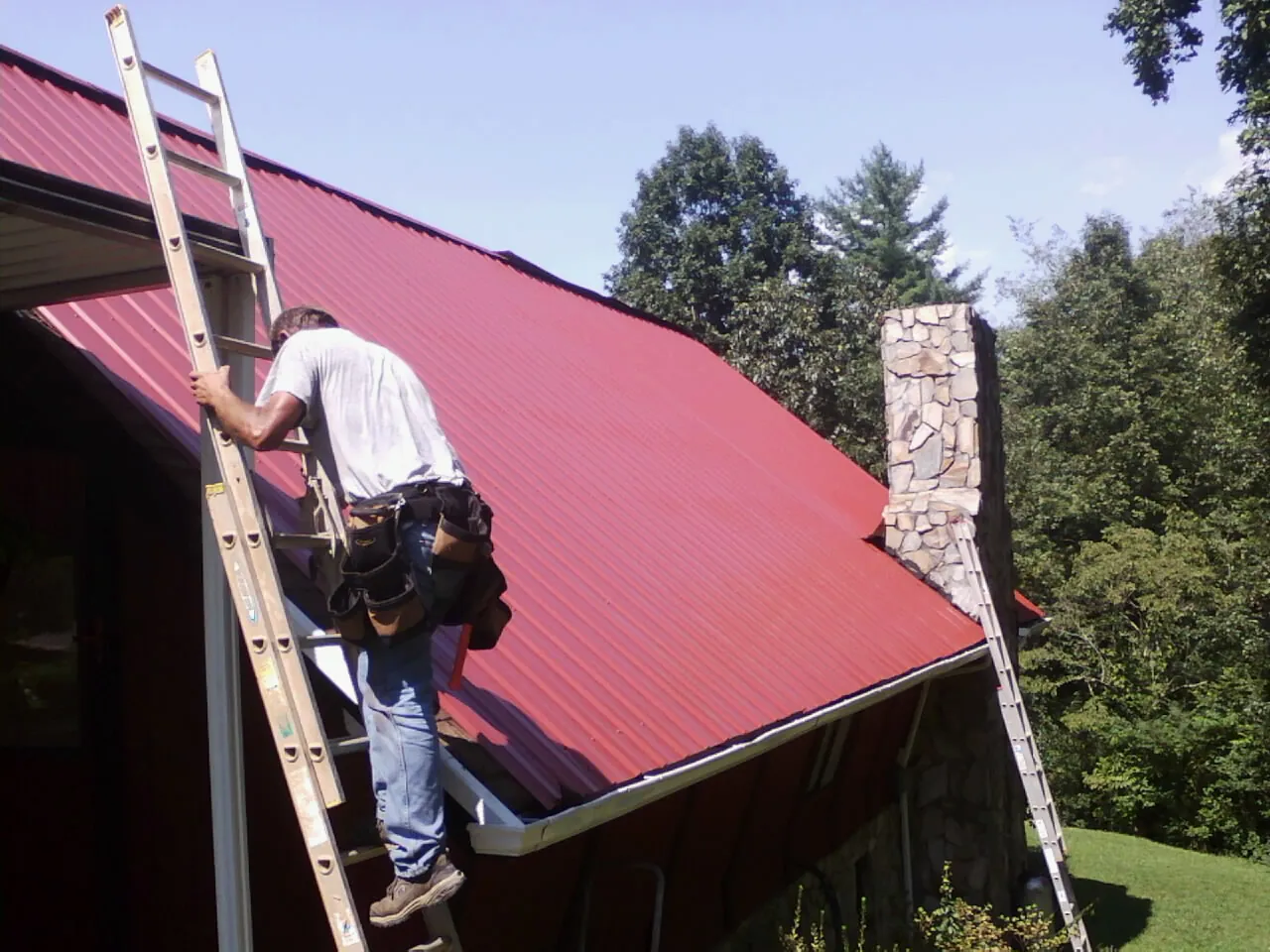 Roofer on a ladder beside a red metal roof and stone chimney — Soffit & Fascia Repair by Roofing Brothers in Belfair